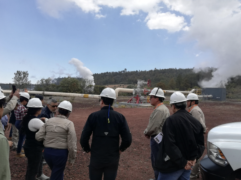 Group of workers in hard hats listening to a briefing near an industrial pipeline with steam plumes in the background.