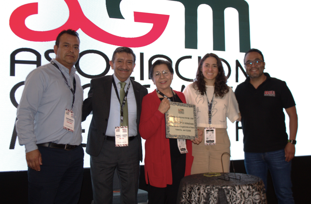 Group of five adults posing on stage, with one woman in a red jacket holding a framed award plaque, backdrop shows a large logo and text.